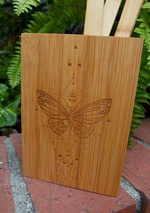 A bamboo panel laser engraved with a detailed butterfly and fine geometric lines, standing on a brick step in front of ferns
