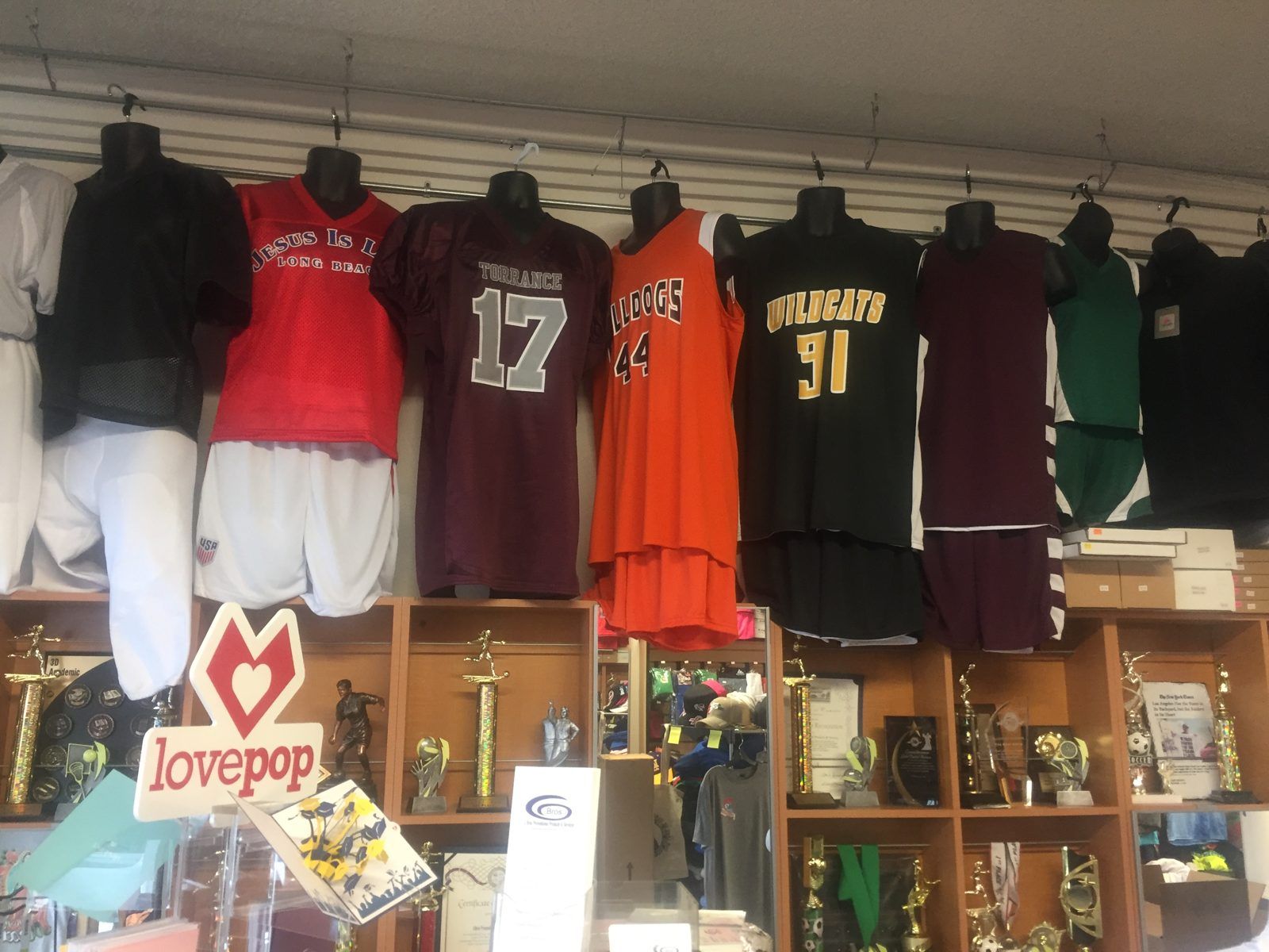 Soccer, football and basketball uniforms with team names and numbers, hanging above the trophy shelves in our shop