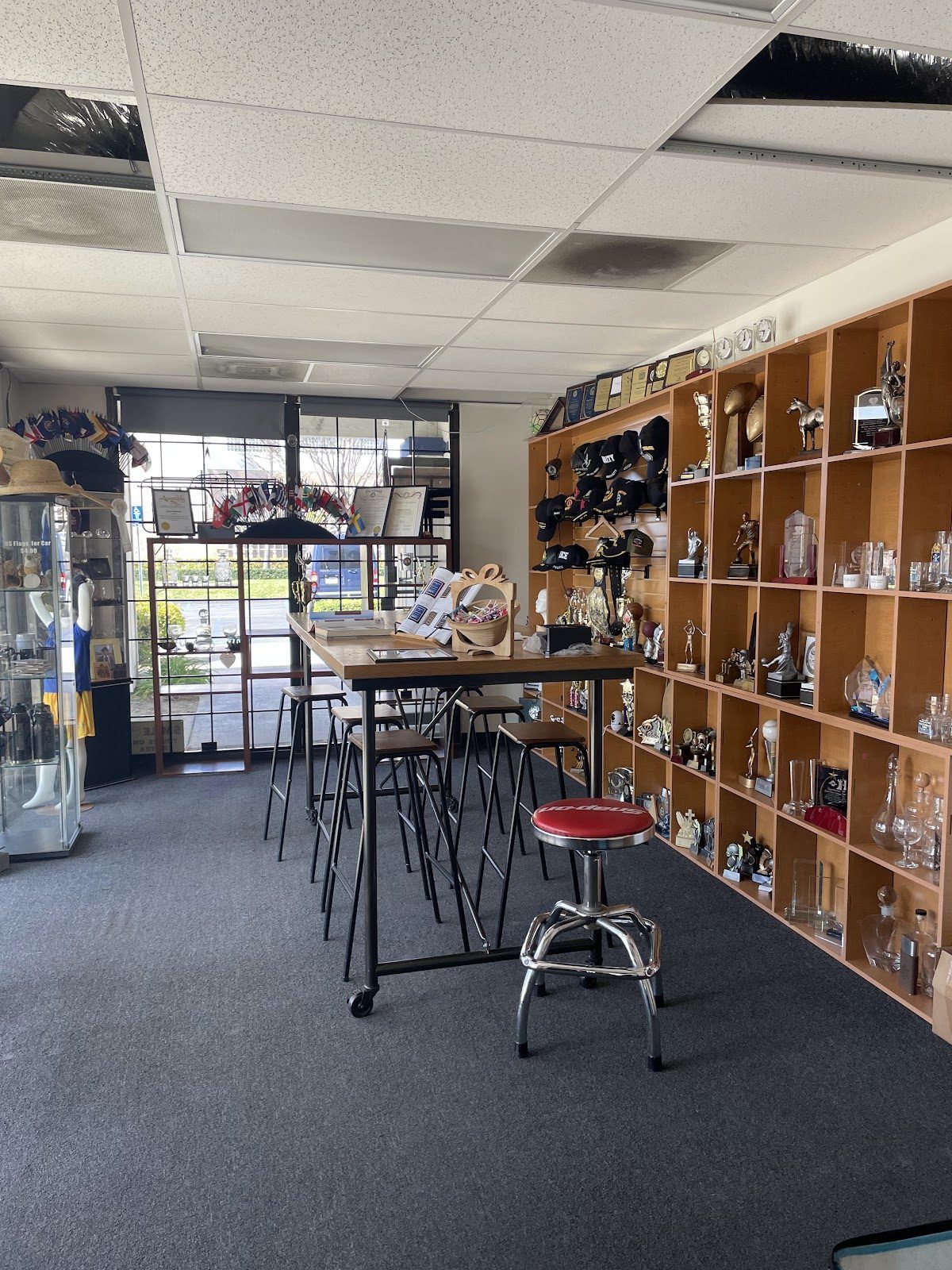 The front room of our shop: a long wooden table with stools, maple cubbies of trophies and caps, and a display case by the front windows