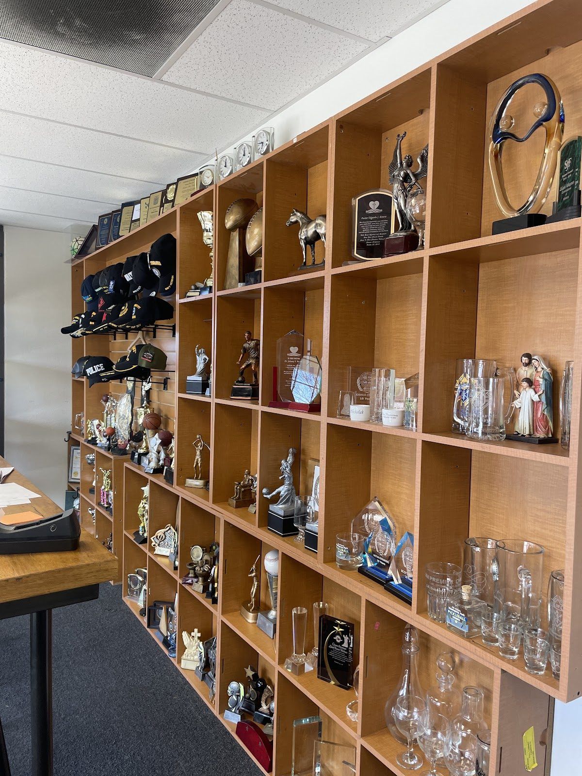 The showroom wall: maple cubbies of sports trophies, crystal and acrylic awards, glass mugs and decanters and a Holy Family statue, with embroidered caps on the end wall