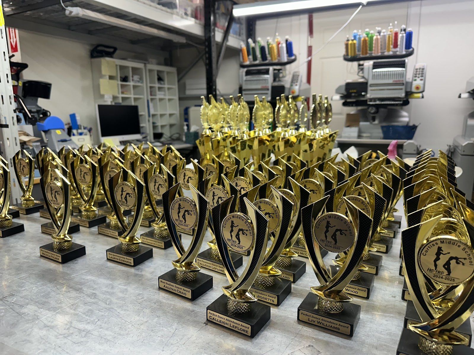 Rows of gold dance trophies for a middle school’s hip hop program, each on a black base with an engraved name plate, and our embroidery machines behind them